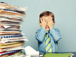 Stacks of worthless stock tax paperwork sit on a desk beside a child in button up shirt and tie holding his face in his hands in frustration.