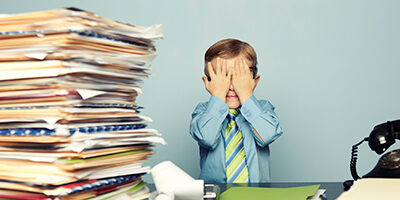 Stacks of worthless stock tax paperwork sit on a desk beside a child in button up shirt and tie holding his face in his hands in frustration.