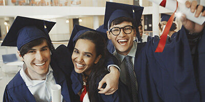 Three jovial students in graduation cap and gown. clutching their diplomas, celebrate the above The Line Education Tax Deduction Reinstatment