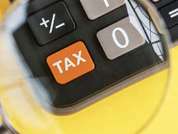 What the April 15 Tax Filing Deadline Extension Means to You. A magnifying glass is held over a calculator, enlarging a button with the word TAX written on it.