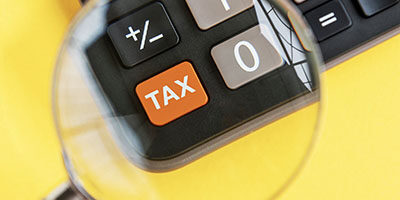 What the April 15 Tax Filing Deadline Extension Means to You. A magnifying glass is held over a calculator, enlarging a button with the word TAX written on it.