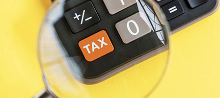 What the April 15 Tax Filing Deadline Extension Means to You. A magnifying glass is held over a calculator, enlarging a button with the word TAX written on it.