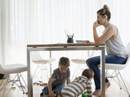 Working from Home During The COVID-19 Outbreak. a woman sits at a kitchen table working on a laptop with two little ones playing underneath her.