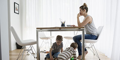 Working from Home During The COVID-19 Outbreak. a woman sits at a kitchen table working on a laptop with two little ones playing underneath her.