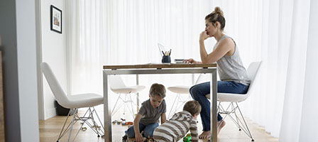 Working from Home During The COVID-19 Outbreak. a woman sits at a kitchen table working on a laptop with two little ones playing underneath her.