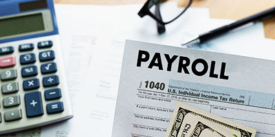 Employers Can Defer Payroll Taxes, show a calculator, PAYROLL sheet and a pair of glasses sitting on a desk.