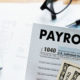 Employers Can Defer Payroll Taxes, show a calculator, PAYROLL sheet and a pair of glasses sitting on a desk.