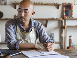 Video: Self-Employed Individuals Now Qualify For Unemployment. A tradesman works on a laptop in his workshop wearing a shop apron