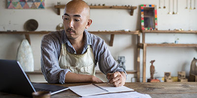 Video: Self-Employed Individuals Now Qualify For Unemployment. A tradesman works on a laptop in his workshop wearing a shop apron