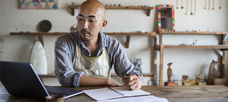 Video: Self-Employed Individuals Now Qualify For Unemployment. A tradesman works on a laptop in his workshop wearing a shop apron