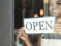 Paycheck Protection Program and Health Care Enhancement Act: What's in It? A business woman flips around a store sign to the OPEN side and smiles, ready for business.