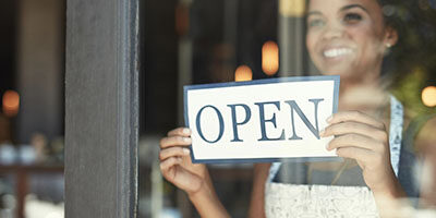 Paycheck Protection Program and Health Care Enhancement Act: What's in It? A business woman flips around a store sign to the OPEN side and smiles, ready for business.