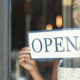 Paycheck Protection Program and Health Care Enhancement Act: What's in It? A business woman flips around a store sign to the OPEN side and smiles, ready for business.