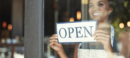 Paycheck Protection Program and Health Care Enhancement Act: What's in It? A business woman flips around a store sign to the OPEN side and smiles, ready for business.