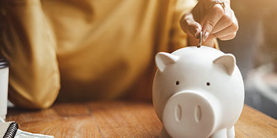 2020 RMD Rollover Relief - A hand puts a penny in a pale pink ceramic piggy bank