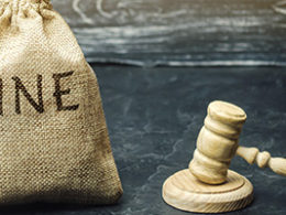 Watch out for tax penalties. A filled burlap sack with the words FINE printed on it, sits next to a wooden gavel on a desk.