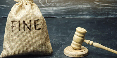 Watch out for tax penalties. A filled burlap sack with the words FINE printed on it, sits next to a wooden gavel on a desk.