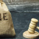 Watch out for tax penalties. A filled burlap sack with the words FINE printed on it, sits next to a wooden gavel on a desk.