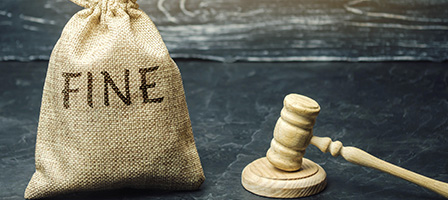 Watch out for tax penalties. A filled burlap sack with the words FINE printed on it, sits next to a wooden gavel on a desk.