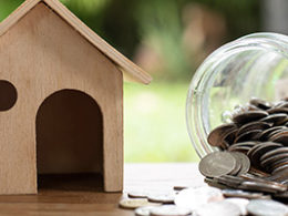 Are You Paying Too Much Interest on Your Home Mortgage? A wooden model of a house sits on a bench beside a jar of coins that is on it side. Coins spill out onto the table top and onto the ground