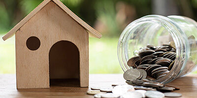 Are You Paying Too Much Interest on Your Home Mortgage? A wooden model of a house sits on a bench beside a jar of coins that is on it side. Coins spill out onto the table top and onto the ground