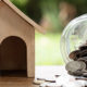 Are You Paying Too Much Interest on Your Home Mortgage? A wooden model of a house sits on a bench beside a jar of coins that is on it side. Coins spill out onto the table top and onto the ground