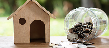 Are You Paying Too Much Interest on Your Home Mortgage? A wooden model of a house sits on a bench beside a jar of coins that is on it side. Coins spill out onto the table top and onto the ground