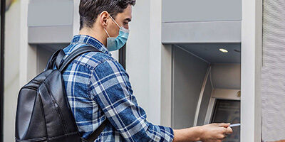 Do You Know Unemployment Benefits Are Taxable? A young man approaches a cash machine wearing a mask due to COVID.