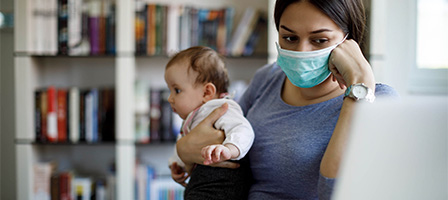 Video: Do You Know That Unemployment Benefits Are Taxable? A woman wearing a mask, holding a baby in her arms while she works at a computer at a public library