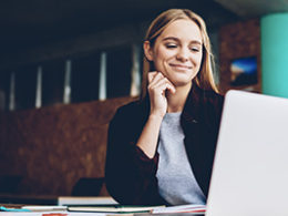 Actually, A Recession Is A Great Time to Launch That New Startup says a woman smiling proudly at her laptop, dressed in business casual clothes.