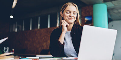 Actually, A Recession Is A Great Time to Launch That New Startup says a woman smiling proudly at her laptop, dressed in business casual clothes.