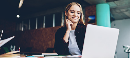 Actually, A Recession Is A Great Time to Launch That New Startup says a woman smiling proudly at her laptop, dressed in business casual clothes.