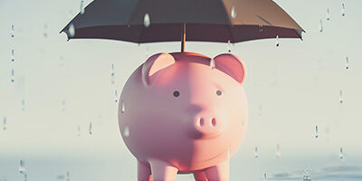 Inflation and Recession: A Double Whammy For Your Finances a piggy bank with an umbrella over it, protecting it from the rain.