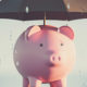 Inflation and Recession: A Double Whammy For Your Finances a piggy bank with an umbrella over it, protecting it from the rain.