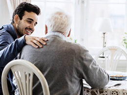 2023 Social Security Cost Of Living Increase Highest In 40 Years A young man puts his arm around an older man while they sit together at a cozy kitchen table.