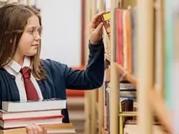 Understanding the Kiddie Tax: a youth holding a stack a books walks down a library aisle reading titles.