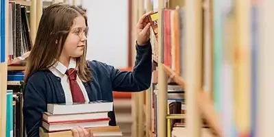 Understanding the Kiddie Tax: a youth holding a stack a books walks down a library aisle reading titles.