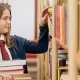 Understanding the Kiddie Tax: a youth holding a stack a books walks down a library aisle reading titles.