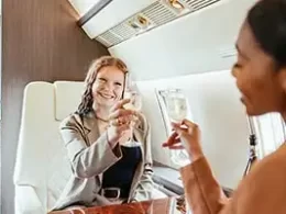 Tax Strategies For The Rich. Two well dressed and impeccably groom attractive ladies click their champagne glasses together while sitting in first class on a private jet.