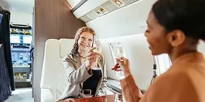 Tax Strategies For The Rich. Two well dressed and impeccably groom attractive ladies click their champagne glasses together while sitting in first class on a private jet.