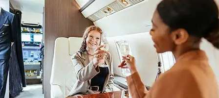 Tax Strategies For The Rich. Two well dressed and impeccably groom attractive ladies click their champagne glasses together while sitting in first class on a private jet.