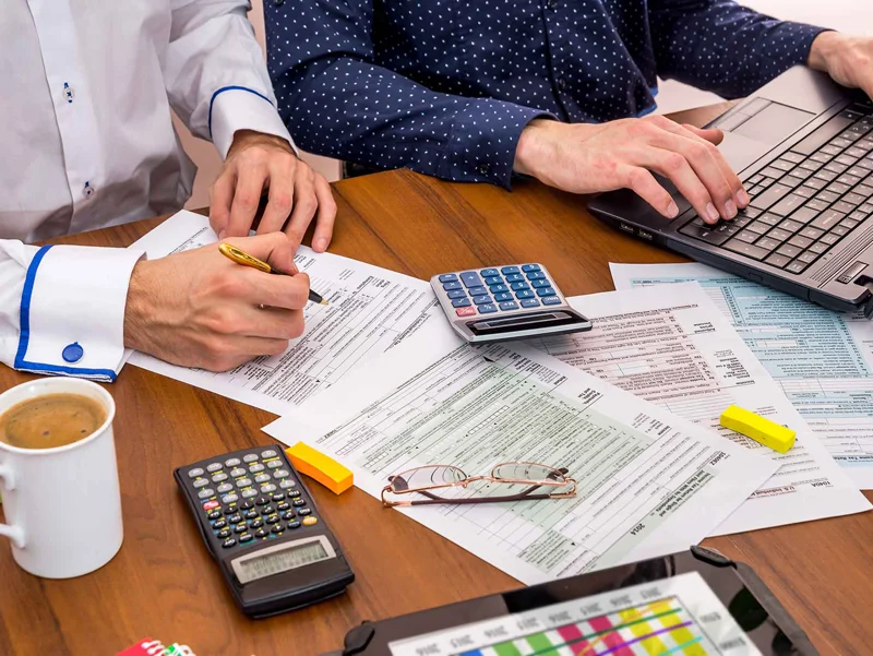 Two people work on tax preparation and planning on a table, surrounded by coffee cups and calculators.