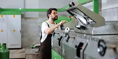 A man in an apron operates a large industrial machine in a factory setting with green accents while considering 100% Bonus Depreciation.