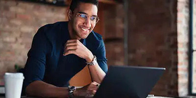 A man in glasses smiles while working on LLC paperwork on a laptop, with a coffee cup nearby.